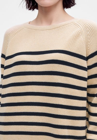 Femme portant un pull en maille beige avec des rayures horizontales noires et un col rond, visible des épaules à la poitrine.