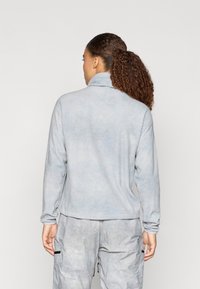 Svetlo modri, mehki flis pulover z visokim ovratnikom, z rahlo teksturo. Obut je z svetlo sivo vzorčasto hlače.