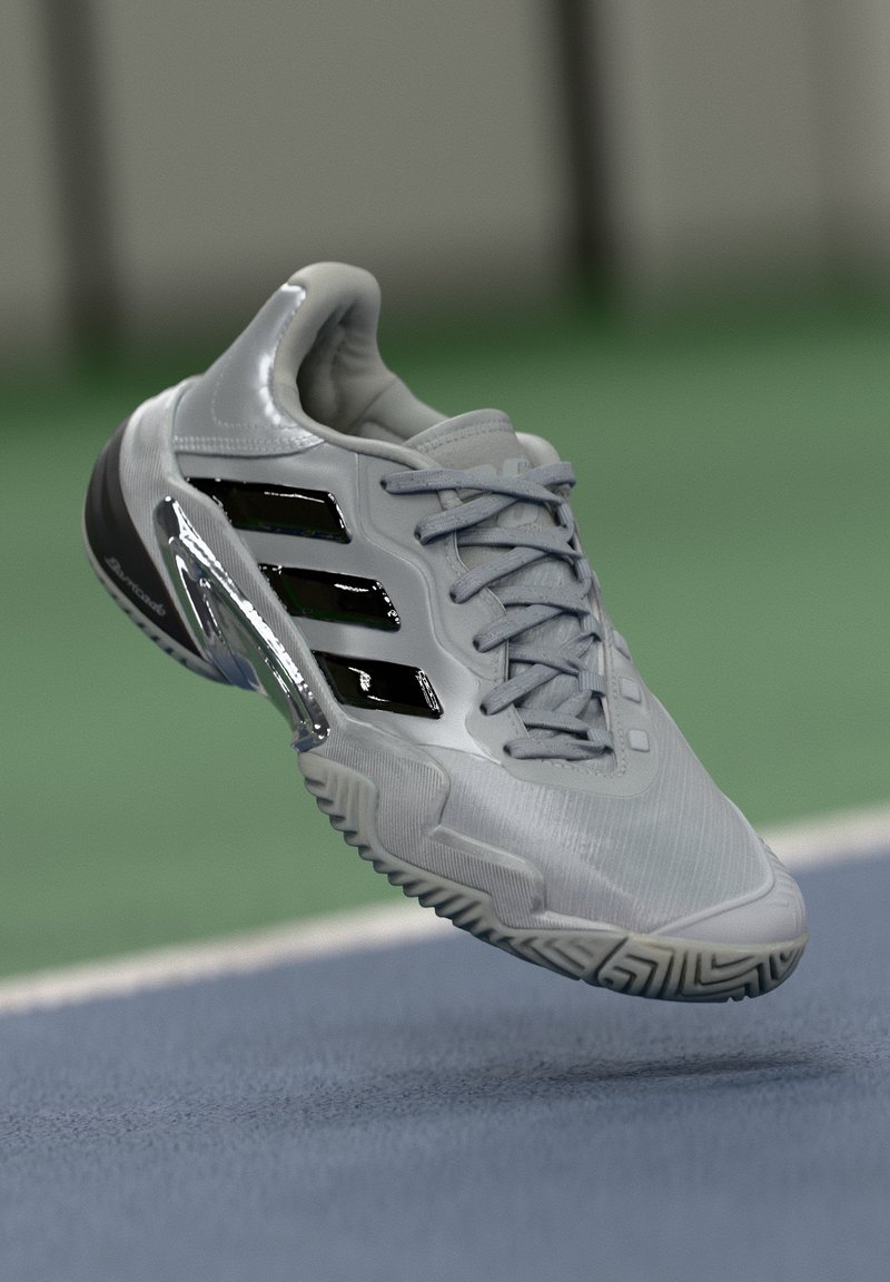 Gray athletic shoe with black stripes and silver heel detail suspended above a tennis court surface.