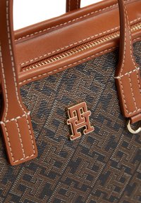 Καφέ και μαύρη τσάντα με μοτίβο, με λαβές από δέρμα ταμπά, λευκή ραφή, χρυστό φερμουάρ και έμβλημα με λογότυπο "TH" σε χρυσό και καφέ.