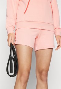 Lys rosa bomullsshorts med en tettsittende design og sideaksenter. Personen holder et svart hoppetau, og viser sporty treningsklær.