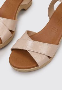 Sandalias cruzadas en color beige con un acabado brillante, plantilla en tono marrón y un bajo tacón de madera. El diseño combina confort y sencillez.