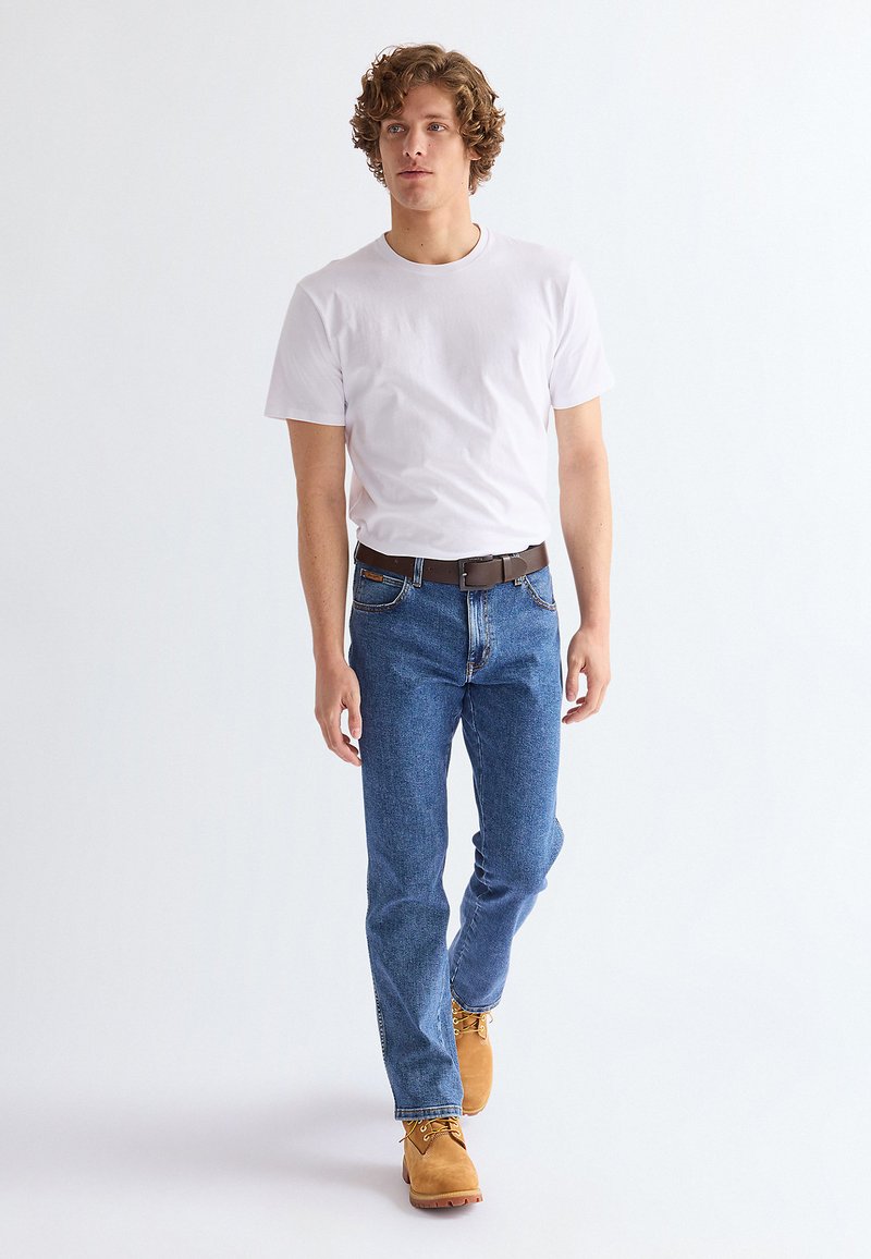 Jeune homme aux cheveux bouclés marchant, portant un t-shirt blanc, un jean bleu, une ceinture marron et des bottes lacées couleur beige sur un fond uni.