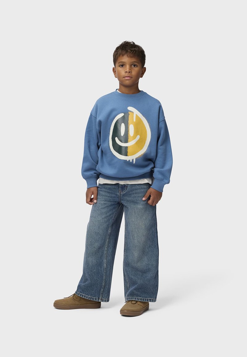 Garçon debout portant un sweat-shirt bleu avec un visage smiley moitié noir, moitié jaune, un jean large et des chaussures marron sur un fond uni.