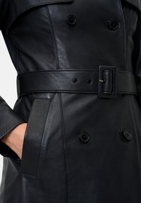Black leather trench coat with double-breasted buttons, belted waist, side pockets, and smooth texture, highlighting stitching details.