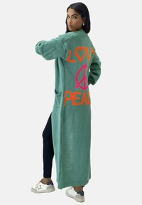 Lång grön kofta i mjuk stickad kvalitet, med ljusorange "LOVE" och rosa "PEACE" bokstäver på ryggen, samt fickor framtill.