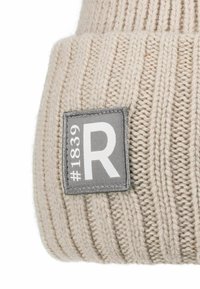 Cream-colored knit beanie with a ribbed texture. Features a gray fabric patch labeled "#1839 R" with white text.