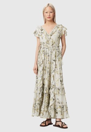 Kobieta ubrana w długą białą sukienkę w kwiaty z falbankami i dopasowaną talią, w połączeniu z czarnymi płaskimi sandałami, stojąca na jednolitym tle.