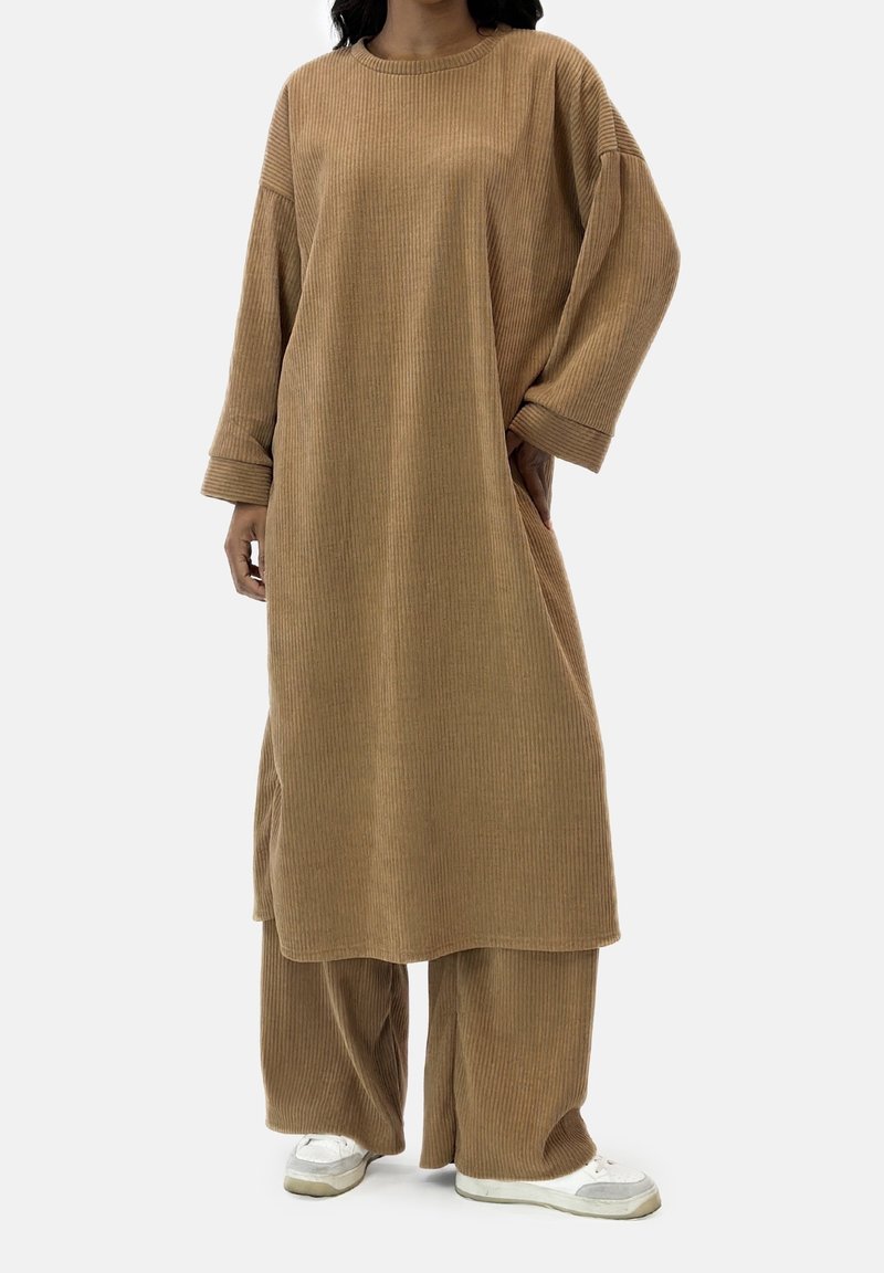 Personne portant une robe longue à manches longues beige côtelée et ample, assortie à un pantalon large de la même couleur, avec des baskets blanches sur un fond blanc.