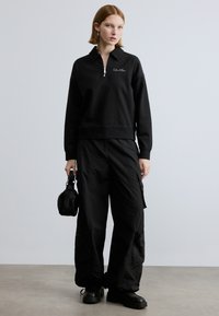 Felpa nera con zip e colletto corto, abbinata a pantaloni cargo neri larghi. Il modello tiene in mano una piccola borsa nera. Tessuto dalla trama liscia.