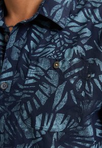 Camicia blu navy con un motivo di foglie testurizzate in aqua. Include una tasca sul petto con un bottone metallico e un piccolo logo sul lato.