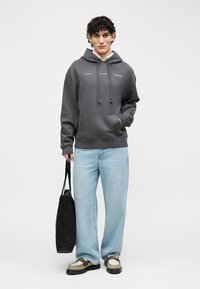 Jaunas vyras dėvi tamsiai pilką džemperį su gobtuvu, šviesiai mėlynas džinsus, dviejų spalvų batus ir laiko juodą rankinę priešais paprastą fono.