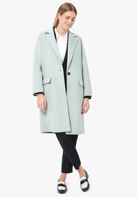 Cappotto in lana verde menta con collo a scialle, chiusura a un bottone, tasche laterali e vestibilità rilassata, indossato sopra pantaloni neri e scarpe.