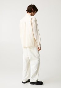 Krém színű, oversized ing hosszú ujjakkal, gombokkal az ujjaknál, off-white, széles szárú nadrággal és fekete cipővel párosítva.