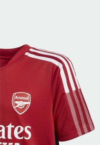 Camiseta de fútbol roja del Arsenal con franjas blancas en los hombros y las mangas, mostrando el escudo del club y texto parcial del patrocinador.