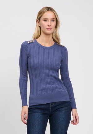 Blonde Frau trägt einen enganliegenden blauen Pullover mit Zopfmuster und goldenen Knöpfen an den Schultern sowie dunkelblaue Jeans, steht vor einem einfarbigen Hintergrund.