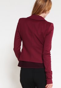 Blazer ajustado burdeos con una textura suave, hombros estructurados y un cuello de muesca simple. Presenta un dobladillo que termina a la altura de la cintura.