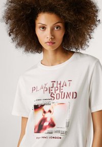 T-shirt bianca in cotone con stampa grafica che include testo e un'immagine di labbra. Vestibilità comoda con scollo a girocollo e tessuto morbido.