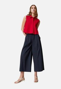 Top senza maniche rosso con bottoni e pieghe, abbinato a pantaloni larghi blu scuro. La modella indossa sandali con tacco bianchi; design minimalista.