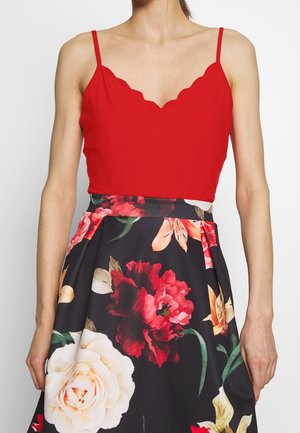 Rød ærmeløs top med bølget halslinje, kombineret med en blomsterprint nederdel med store blomster i rød, hvid og gul på en sort baggrund.