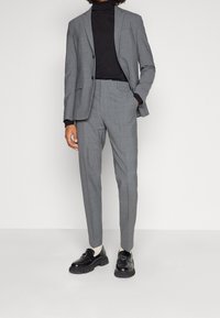 Persona che indossa un completo grigio con pantaloni su misura, dolcevita nero, calze bianche e mocassini neri, in piedi contro uno sfondo bianco semplice.