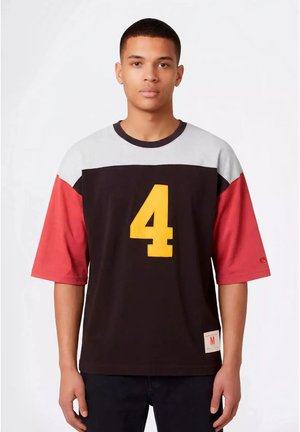 Un jeune homme se tient face à l'avant, portant une chemise noire, blanche et rouge avec un grand numéro 4 jaune sur le devant et un pantalon noir.