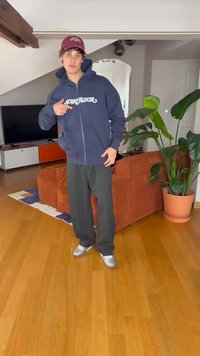 Námořnická zipová mikina s logem "Pescador", šedé kalhoty volného střihu a stříbrné tenisky. Místnost má dřevěnou podlahu a rostlinu v květináči.