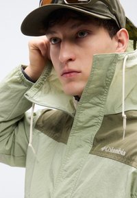 Light green waterproof jacket with a dark green upper panel. Prominent drawstrings at the collar, featuring the Columbia logo on the chest.