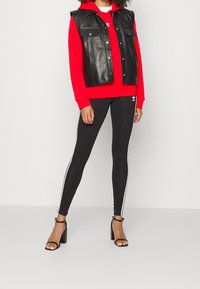 Gilet di pelle nera sopra un hoodie rosso, abbinato a leggings neri con strisce bianche ai lati e sandali con tacco neri.