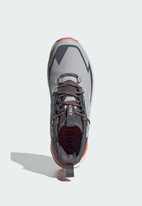 Szare buty sportowe z teksturowaną cholewą, czarnymi i srebrnymi akcentami, pomarańczową podeszwą, odblaskowymi detalami oraz systemem sznurowania zapewniającym pewne dopasowanie.