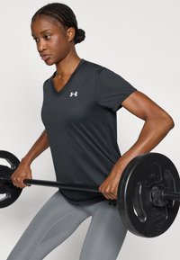 Haut d'entraînement noir à manches courtes et col en V avec logo Under Armour, associé à des leggings gris ajustés, en train de lever une barre.