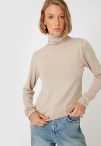 Pull-over beige à col roulé en tissu côtelé, avec des manches longues et une silhouette ajustée, associé à un jean en denim bleu clair.