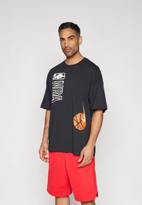 Μαύρο κοντομάνικο μπλουζάκι με λογότυπο NBA και πορτοκαλί γραφικό με μπάλα του μπάσκετ· συνδυασμένο με κόκκινα αθλητικά σορτς· βαμβακερό, άνετη γραμμή.