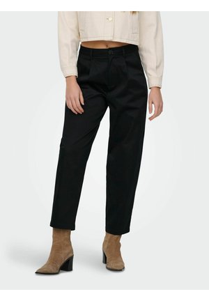 Nő fekete, magas derekú nadrágot, bézs, rövidített dzsekit és világosbarna, nubuk bokacsizmát visel blokk sarkakkal, fehér háttér előtt áll.