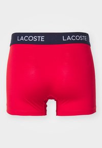 Piros férfi boxeralsó, kék elasztikus derékkal, amelyen fehér "LACOSTE" felirat található. Simító, rugalmas anyagból készült, szoros illeszkedéssel.