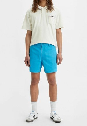 Person iført cremefarvet polo-shirt med korte ærmer, lyseblå shorts, hvide mellemlange sokker og hvide sneakers med sorte striber, der står mod en ensfarvet baggrund.