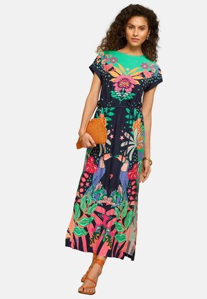 Kvinde iført en livlig kjole med blomstermønster og tukanprint, der holder en orange flettet clutch og har på sig orange sandaler med stropper.