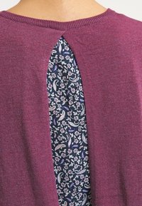 Fioletowy sweter z dzianiny z wykrojem na plecach, ozdobiony ciemnym wzorem kwiatowym, w tym paisley i kwiatami, na kontrastowym tle.