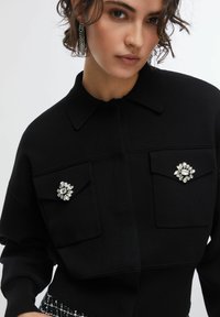 Top nero in maglia con colletto, due tasche frontali e decorazioni gioiello sulle tasche. Tessuto liscio e design aderente.