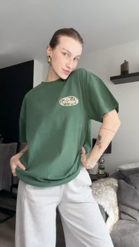 T-shirt verde de algodão com mangas curtas e decote redondo, apresentando um logótipo estampado no peito. Usado com calças de fato cinza claro.