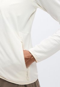 Φωτεινό μπεζ μακρυμάνικο πουλόβερ με υφασμένο, ριγωτό σχέδιο, που διαθέτει τσέπη με φερμουάρ και αντίθετες μανσέτες. Το υλικό φαίνεται μαλακό και διαπνέον.