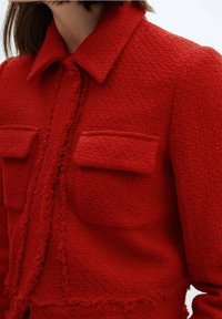 Red tweed jacket featuring a collar, front pockets, and a fringed hem. The texture is textured and slightly nubby, with a structured design.