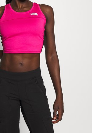 Vrouw draagt een roze cropped tanktop met het logo van The North Face en zwarte broek met ritssluiting en zakken, staand tegen een effen achtergrond.