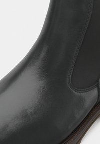 Bota de cano curto em couro preto com acabamento brilhante, painel lateral elástico, bico arredondado e um salto baixo empilhado. Textura lisa, costura mínima.