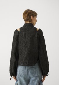 Black textured blouse with a structured collar, open shoulder design, and voluminous sleeves, paired with light blue denim.