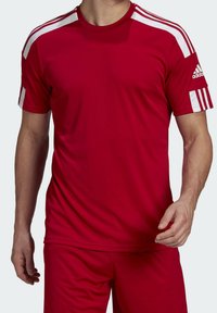 Punainen urheilupaita valkoisilla olkapääraidoilla, valmistettu sileästä kankaasta. Sisältää Adidas-logon hihassa ja on yhdistetty matching-sortseihin.