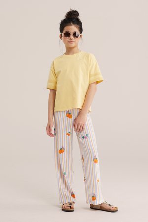 Tüdruk, kes kannab kollast lühikeste varrukatega särki, triibulisi pükse apelsinimotiividega, lillikujulisi päikeseprille ja sandaale, seisab lõõgastunud poosis.