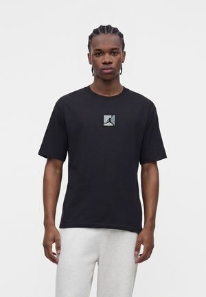Ung mand med flettet hår iført sort kortærmet t-shirt med et lille Jordan-logo og lysegrå bukser mod en ensfarvet baggrund.