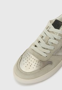 Sneaker grigio chiaro con tomaia in suede e punta in pelle metallizzata. Presenta dettagli perforati, lacci piatti e una toppa logo nera.