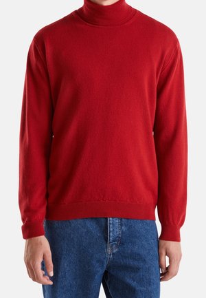 Persona che indossa un maglione a collo alto a maniche lunghe rosso e jeans blu, mostrata dalla metà del collo fino ai fianchi contro uno sfondo semplice.